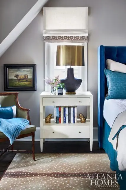 Bedroom with a blue tufted headboard, a green armchair with a blue throw, a white nightstand with books and a lamp, and a fawn-print rug.