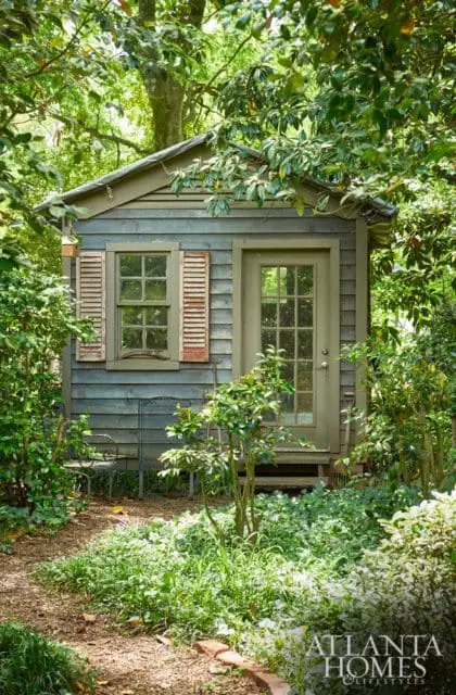 Small weathered wooden shed with green door and window with distressed shutters, surrounded by lush green foliage and trees.