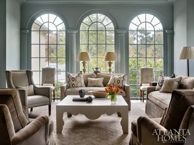 Living room with three arched windows overlooking trees, featuring neutral-toned sofas and armchairs, a white coffee table, and a patterned rug.