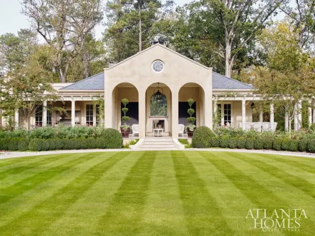 Cream stucco house with arched entryway, slate roof, and covered porches. Lush landscaping and manicured lawn in front.