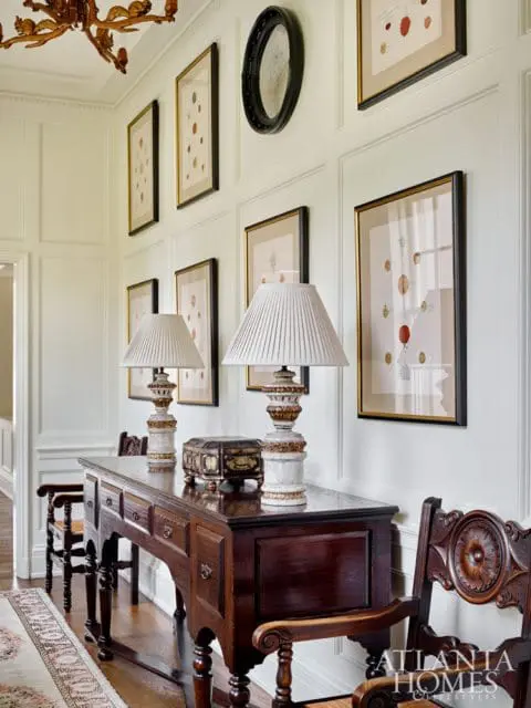 Hallway with paneled walls, a dark wood console table with two lamps and a decorative box, framed botanical prints, and ornate wooden chairs.
