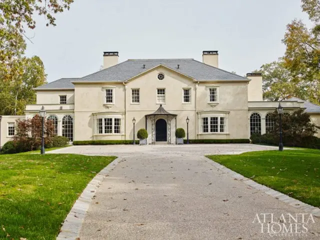 Large cream-colored stucco house with a gray slate roof, multiple bay windows, arched windows, and a covered entryway. A gravel driveway leads to the house, flanked by green lawns and mature trees.