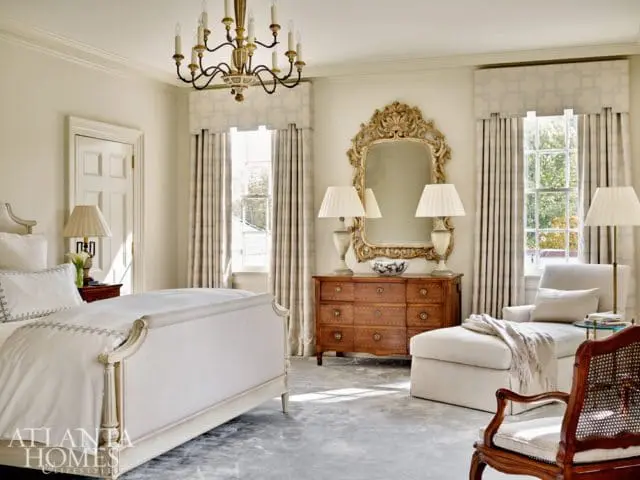 Cream bedroom with a tufted bed, ornate gold mirror above a wooden dresser, and a chaise lounge with a throw blanket.