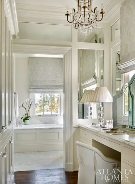 White dressing room with a vanity, upholstered chair, mirrored walls, and a crystal chandelier. A window with a roman shade looks into a marble-clad bathroom.