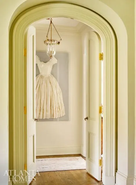 Cream-colored dress displayed in a framed artwork hangs on a cream wall in a hallway with an arched doorway, brass-hinged doors, and a glass lantern chandelier.