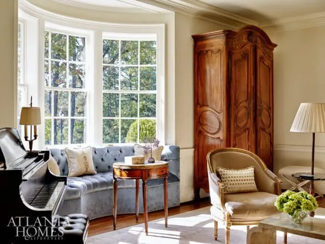 Living room with a black grand piano, tufted blue curved sofa, antique wooden armoire, and a French-style armchair with a floral arrangement.
