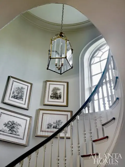 Curved staircase with white balusters and dark handrail, adorned with four framed botanical prints and a brass and glass lantern chandelier.