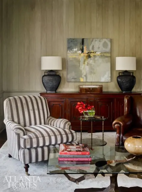 Living room with striped armchair, leather wingback chair, dark wood credenza, abstract art, and glass coffee table with books.
