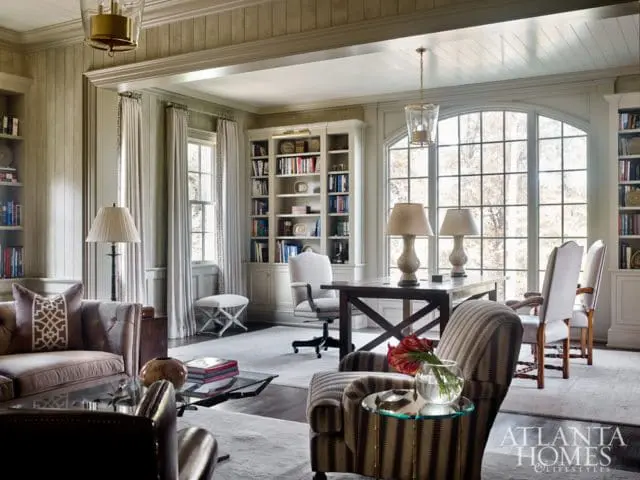 Study with built-in bookshelves, tufted sofa, patterned armchair, and large arched window with a view of trees.