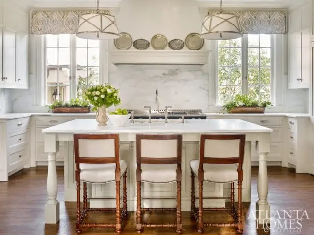 White kitchen with marble backsplash, large island with three upholstered stools, and two pendant lights.
