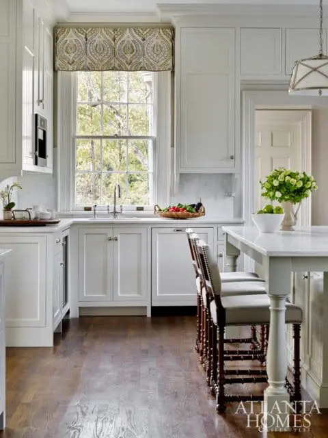 White kitchen with marble countertops, paneled cabinets, and a large window overlooking trees. Bar stools with dark wood frames and upholstered seats are at a marble-topped island.