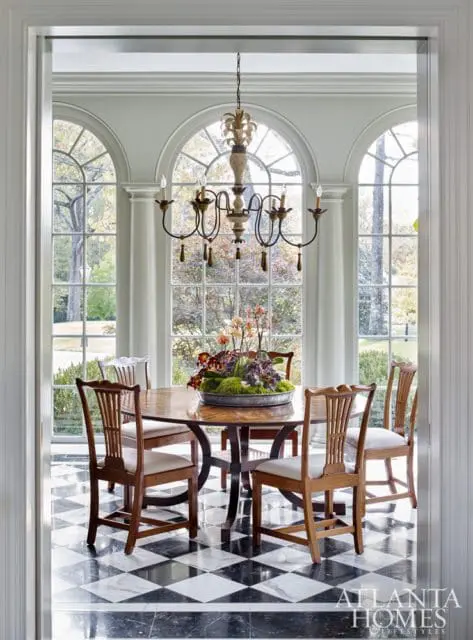 Dining room with a round wood table and chairs, set against a wall of arched windows. Features a black and white marble tile floor and a decorative chandelier.