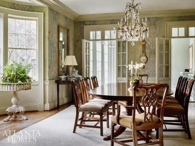Elegant dining room with a large oval wooden table, upholstered chairs, crystal chandelier, patterned wallpaper, and a grandfather clock.