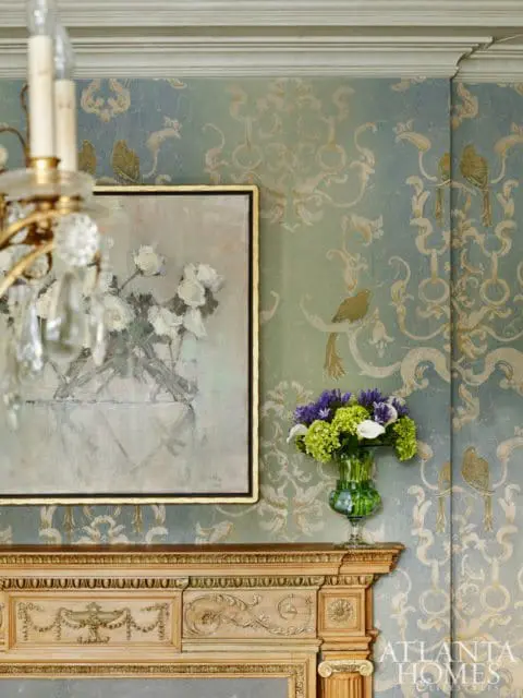 Ornate wooden mantelpiece below a painting of white roses and a vase of purple and green flowers, set against blue and green damask wallpaper.