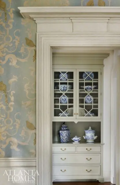 Built-in light gray cabinet with glass doors displaying blue and white porcelain dishes, and drawers with brass hardware. Blue and white ginger jars and a teapot sit on top of the drawers.