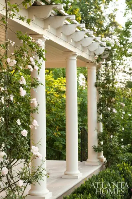 White pergola with decorative trim supported by fluted columns, adorned with climbing pink roses. Lush greenery and trees in the background.
