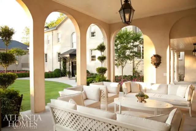 Covered patio with arched openings, featuring white upholstered sofas and chairs, a white oval coffee table, and a view of a manicured lawn and house.