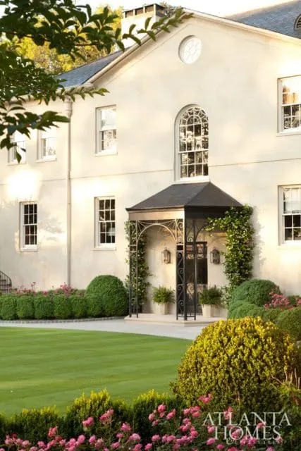 Cream stucco house with arched window above black wrought iron porch, manicured hedges, and blooming pink flowers in the foreground.
