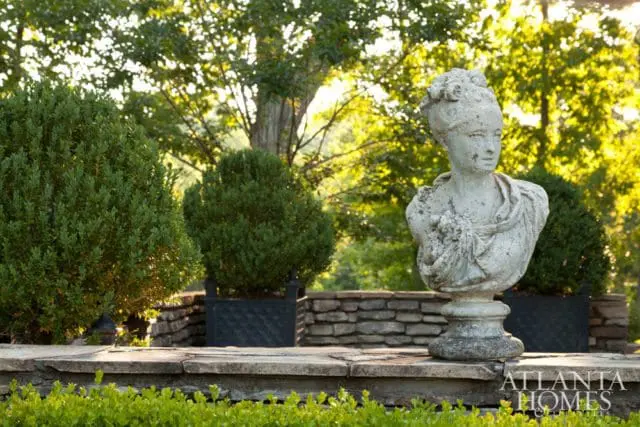 Stone bust of a woman with an ornate hairstyle stands on a stone ledge in a garden with manicured boxwood bushes and trees.