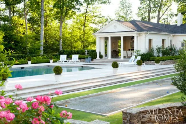 White pool house with columns, pool, bocce court, and manicured landscaping with pink roses in the foreground.