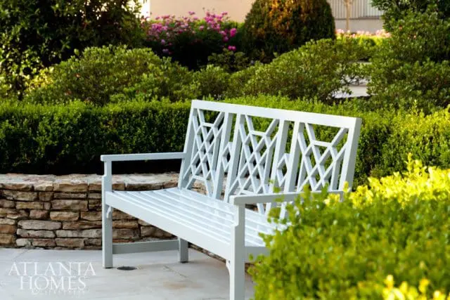White lattice-back bench in a lush garden with manicured hedges, flowering bushes, and a stone retaining wall.