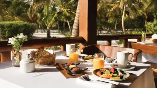 Table set for breakfast with fruit, bread, juice, and coffee on a white tablecloth, with palm trees visible in the background.