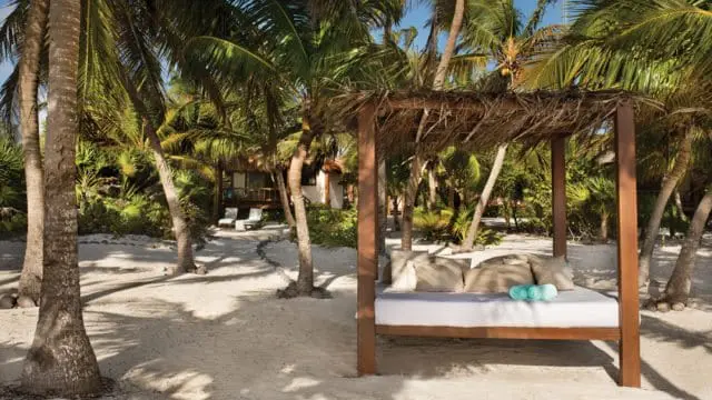 Beach cabana with a daybed, pillows, and rolled towels, surrounded by palm trees and lush greenery.