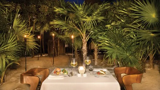 Table set for two with white tablecloth, wine glasses, and plates of food, surrounded by palm trees and lit by tiki torches at dusk.