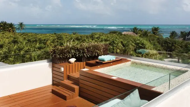 Rooftop jacuzzi with wooden deck and glass railing overlooking lush tropical foliage and the ocean