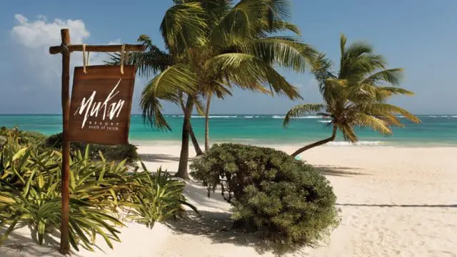 Wooden sign for Mukam Resort south of Tulum on a sandy beach with palm trees and turquoise ocean in the background.