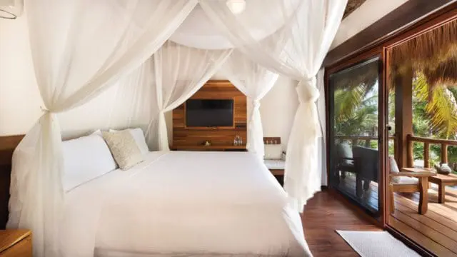 Bedroom with a white canopy bed draped in sheer white fabric, a wooden accent wall with a mounted television, and a sliding glass door opening to a tropical balcony.