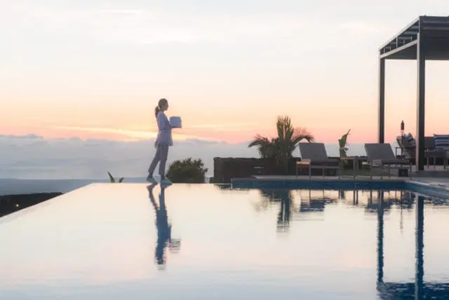 Woman carrying towels walks beside an infinity pool at sunset, with lounge chairs and a cabana visible.
