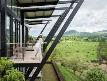 Modern building with black metal beams and glass windows overlooking a lush green landscape with rolling hills under a cloudy sky.