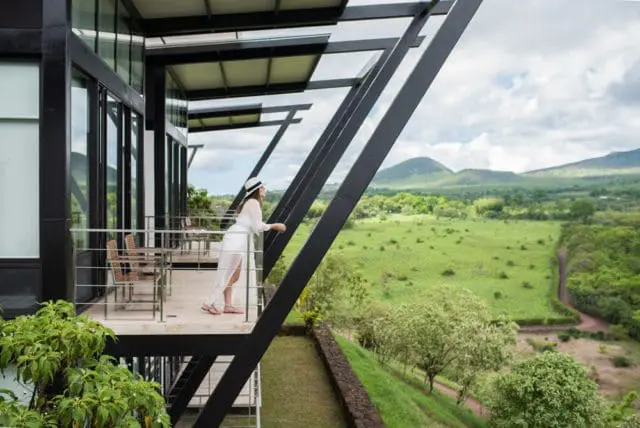 Modern building with black metal beams and glass windows overlooking a lush green landscape with rolling hills under a cloudy sky.