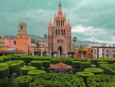 Ornate pink stone church with spires and bell tower, surrounded by manicured green hedges and a gazebo in a town square.