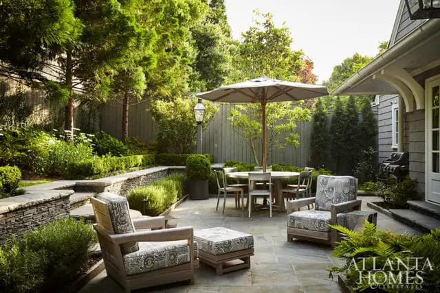 Stone patio with outdoor dining set, chairs, and ottoman surrounded by lush greenery and a wooden fence.
