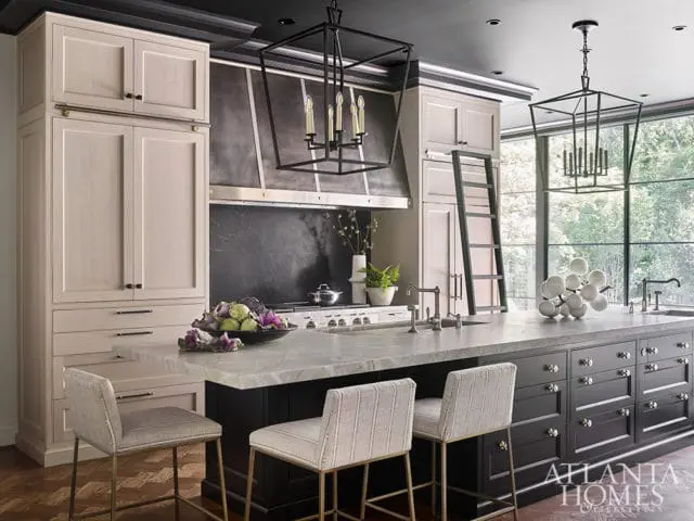Modern kitchen with light wood cabinetry, dark island, marble countertops, and large windows overlooking trees.