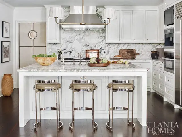 White kitchen with marble island, marble backsplash, stainless steel appliances, and three chrome bar stools with tan upholstery.