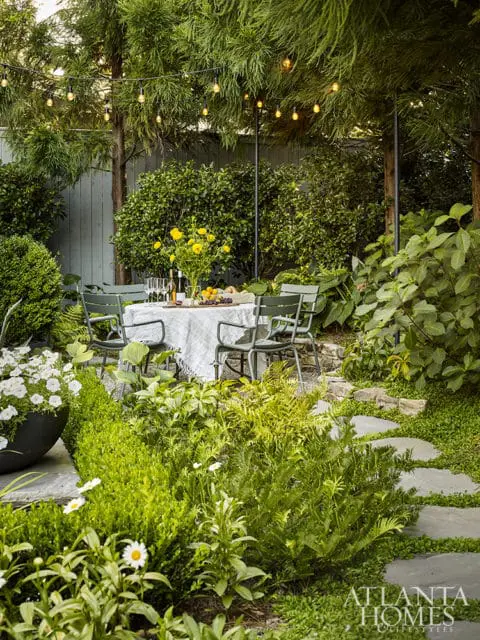 Outdoor dining area with a table set for a meal, surrounded by lush greenery and string lights overhead. Stone pathway leads through the garden.
