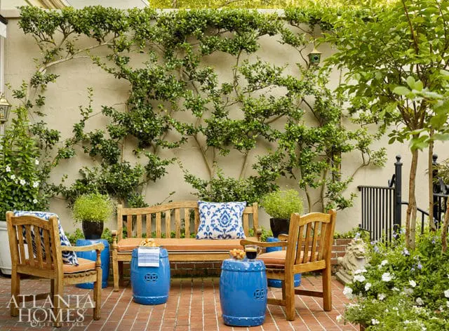 Wooden bench and chairs on a brick patio, adorned with blue ceramic garden stools and patterned cushions, set against a stucco wall covered in climbing ivy.