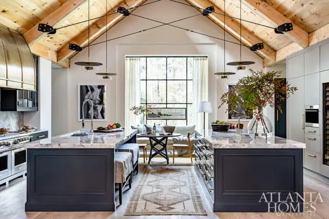 Spacious kitchen with vaulted wood-beamed ceiling, two marble-topped islands with dark gray bases, and a large window overlooking a wooded area.