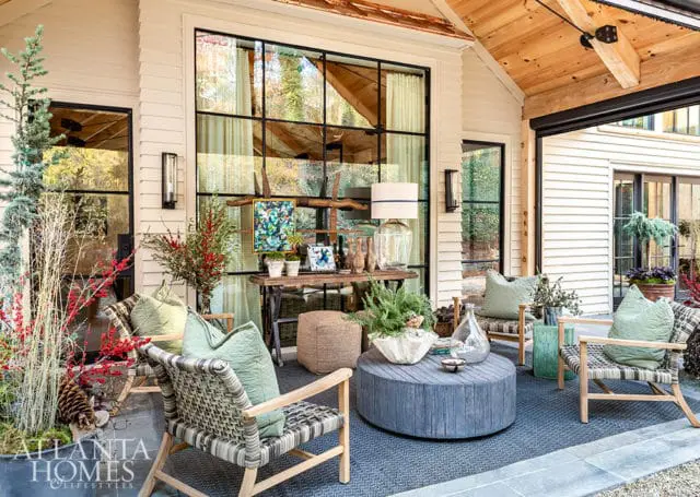 Covered porch with light-colored siding, wood-paneled ceiling, and large black-framed windows. Furnished with woven chairs, a blue circular coffee table, and decorative plants.