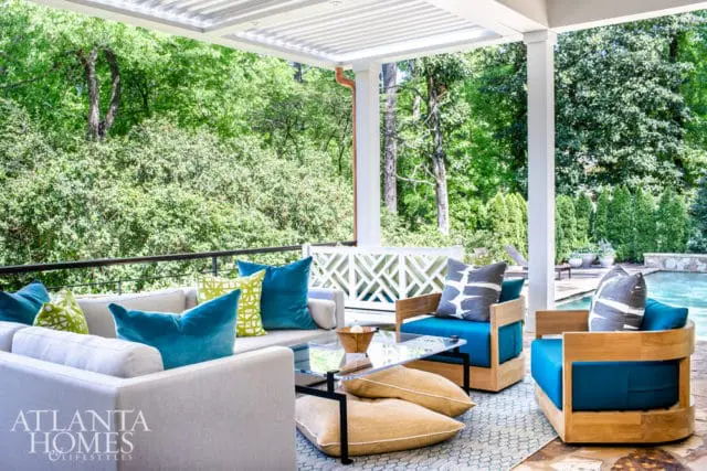 Covered patio with light-colored sectional sofa and wooden chairs with teal cushions, accented by teal and lime green pillows. A glass-top coffee table sits on a patterned rug, with a lush green landscape and pool visible in the background.