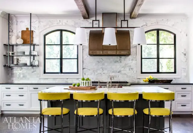 Modern kitchen with white marble backsplash, arched windows, and a large island with yellow velvet bar stools.