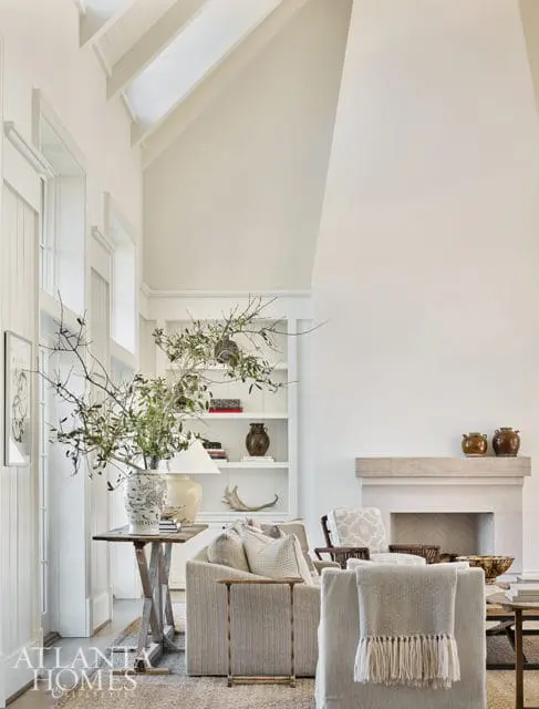 Living room with vaulted ceiling, white walls, built-in white bookshelves, and a stone fireplace. Furnishings include a textured sofa with neutral throw pillows and a fringed blanket.