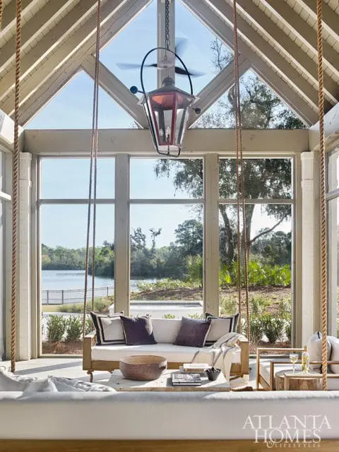 Sunroom with a large window overlooking a lake, featuring a wooden swing sofa with plush cushions, a rustic coffee table, and a hanging lantern with a ceiling fan.