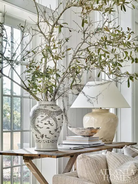 Console table with a large ceramic vase filled with branches, a lamp, and stacked books. A sofa with textured pillows is in the foreground.