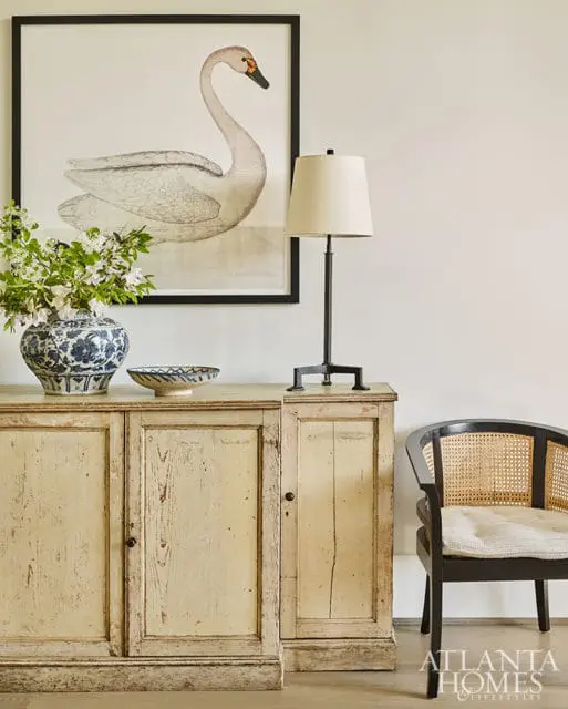 Distressed cream sideboard with a framed swan print above, a blue and white vase with flowers, a lamp, and a black cane-backed chair