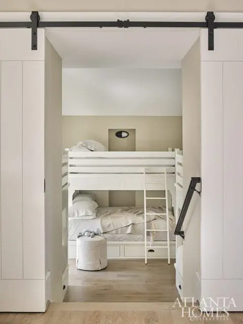 Bunk room with white bunk beds, neutral bedding, a striped ottoman, and a black barn door track overhead.