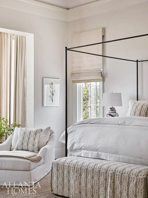 Bedroom with a white canopy bed, white bedding, a white chaise lounge with patterned pillows, and a patterned ottoman.
